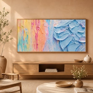 Peut inclure: Tableau abstrait encadré de bois. L'œuvre présente un design texturé et coloré avec des nuances de jaune, de rose, d'orange et de bleu. Le tableau est exposé au-dessus d'un meuble en bois dans une pièce lumineuse.