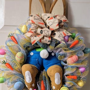 Może przedstawiać: Wieniec wielkanocny z motywem królika. Wieniec ma siatkową podstawę w kolorze żółtym, niebieskim i białym, ozdobioną kolorowymi jajkami i marchewkami. Zawiera uszy królika, kokardę ze wzorami królików i marchewek oraz nogi w dżinsach z puszystym ogonem.