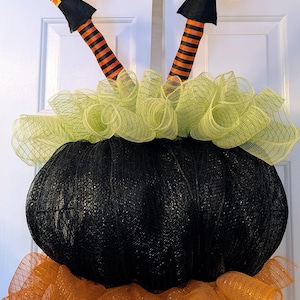 May include: A decorative wreath featuring a large, black, mesh-textured cauldron shape. Two stuffed legs with black and orange striped stockings and black boots extend upward from the top. Lime green mesh ruffles the top, and orange mesh ruffles the base.