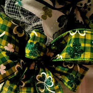 May include: Close-up of a St. Patrick's Day wreath featuring green and yellow plaid ribbons. The ribbons are decorated with shamrocks and glitter. A white ribbon with a green truck graphic is also visible.