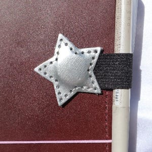May include: A silver star-shaped pen holder attached to a black elastic band, secured to a dark maroon notebook cover. The star features visible stitching along its edges, and a light-colored pen is inserted into the elastic loop on the side.