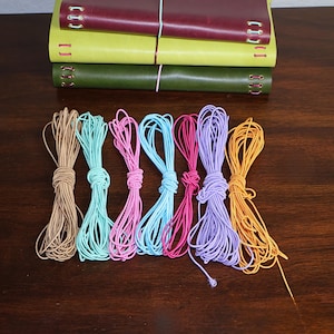 May include: Three rectangular journals in burgundy, yellow, and olive green are stacked horizontally. Below them are seven coiled bundles of thin elastic cord in tan, turquoise, pink, light blue, magenta, lavender, and orange colors.