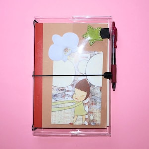 May include: A clear plastic notebook cover holding a tan journal with a red spine. The cover features a cartoon character illustration, a white flower cutout, and a green star sticker. A black elastic band secures the journal, holding a red pen.