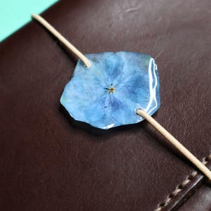 May include: A dark brown leather journal secured with a light beige elastic cord. A single, pressed blue hydrangea flower encased in a clear, glossy resin disc is threaded onto the cord, serving as a decorative closure for the notebook cover.