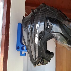 May include: A black motorcycle helmet with white lettering, mounted on a blue wall bracket. The helmet has a clear visor and a carbon fiber pattern. The bracket is attached to a white wall.