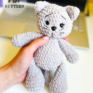May include: A handmade gray crochet cat plushie with black eyes and whiskers. The plushie is held in a hand. The text "PDF PATTERN" is in a circle in the upper left corner. The cat has a cute, slightly sad expression.