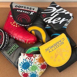 May include: A selection of golf club head covers in diverse colours and designs. Features covers with the "Odyssey Metal-X" and "Scotty Cameron" logos, alongside covers with embroidered patterns and text.