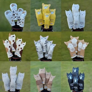 May include: A collection of golf club head covers featuring various cat designs. The covers are in different colors, including white, yellow, brown, and black. Some covers have cat faces, while others have cat paw prints. The text "Dully Na Cat" is printed on the covers.