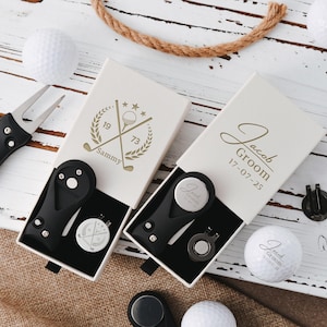 Pode incluir: Duas caixas de presente abertas, cor marfim, com acessórios de golfe, incluindo uma ferramenta de reparação de relva e um marcador de bola. Uma caixa é personalizada com "Sammy" e a outra com "Jacob Groom 17-07-25". Bolas de golfe e um tee também são visíveis.