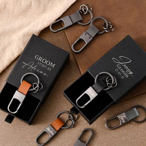 Op de afbeelding: Gepersonaliseerde sleutelhangers met zwarte en bruine leren bandjes en zilverkleurige hardware. Sommige sleutelhangers zitten in zwarte geschenkdozen met het woord "Groom" en een datum erop. De sleutelhangers liggen op een bruin oppervlak.