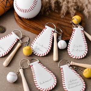 May include: Baseball-themed keychains with white leather tags, red stitching, and miniature baseball bats. Some keychains feature yellow or white baseballs. Text on the tags includes phrases like "Every strike, it's all my goal."
