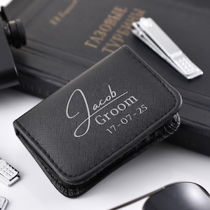 Op de afbeelding: Een zwart leren etui met de naam "Jacob", het woord "Groom" en de datum "17-07-25" in zilveren letters. De etui ligt naast een zwarte fles, een boek, nagelknippers en een zonnebril.