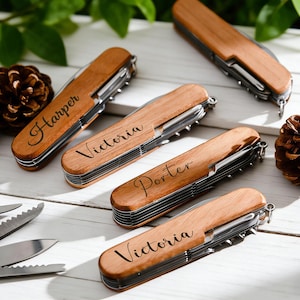 May include: Several personalized wooden pocket knives with stainless steel tools are displayed on a white surface. Each knife has a name engraved on the wooden handle. Pine cones and greenery are in the background.