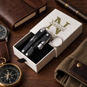 May include: A black multi-tool keyring with a silver ring and the name Ashley engraved on the handle, presented in a white rectangular box featuring the name Natalie printed in large green letters on the top lid with a slide-out design.