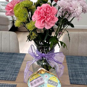May include: A clear glass vase holds a vibrant bouquet of pink carnations, green chrysanthemums, and light purple flowers. A decorative seashell-shaped dish with Easter egg designs sits on a wooden table, adorned with a purple ribbon.