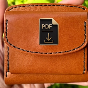 Könnte beinhalten: Ein quadratisches, orangefarbenes Lederportemonnaie mit schwarzen Nähten und einem Klappenverschluss. Auf der Klappe befindet sich ein schwarzes und goldenes PDF-Symbol. Das Portemonnaie wird in einer Hand vor einem verschwommenen grünen Hintergrund gehalten.