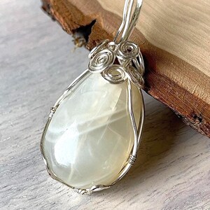 May include: A pear-shaped pendant with a milky white stone, encased in a silver wire frame. The bail features decorative silver swirls. The pendant is displayed on a wooden surface.