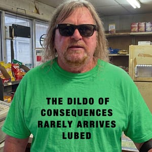 May include: A person wearing a green t-shirt with the text "THE DILDO OF CONSEQUENCES RARELY ARRIVES LUBED" in black. The person is wearing sunglasses and is in a store setting with snacks and a cash register visible.