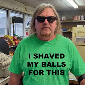May include: A man wearing sunglasses and a green t-shirt with the text "I SHAVED MY BALLS FOR THIS". The man is standing in a store with shelves of snacks and a cash register.