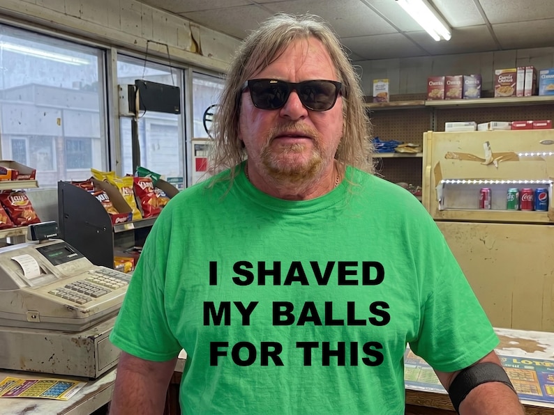 May include: A man wearing sunglasses and a green t-shirt with the text "I SHAVED MY BALLS FOR THIS". The man is standing in a store with shelves of snacks and a cash register.