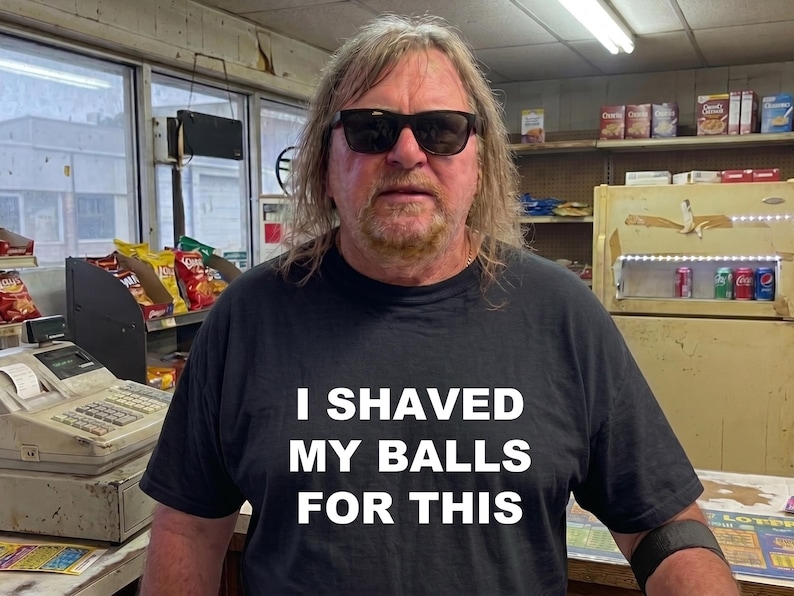 May include: A person wearing a black t-shirt with the text "I shaved my balls for this." The person is wearing sunglasses and standing in a convenience store setting.