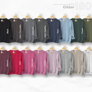 May include: A display of Gildan 18000 crewneck sweatshirts in various colors, including black, navy, and light blue. The sweatshirts are hanging on wooden hangers. The available colors are: Black, Dark Chocolate, Charcoal, Dark Heather, Navy, Light Blue, Forest, Military Green, Maroon, Red, Heliconia, Azalea, Light Pink, Sport Grey, Sand, and White.