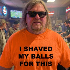 May include: An orange t-shirt with the text "I SHAVED MY BALLS FOR THIS" in black block letters. The person wearing the shirt is in a bar setting, with other people and neon signs in the background.