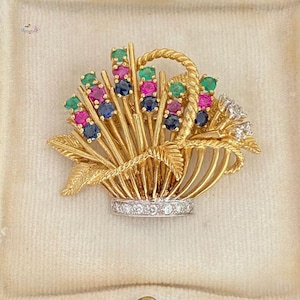 May include: A gold-toned brooch in the shape of a basket overflowing with colorful gemstones. The basket features a braided handle and is adorned with emerald, ruby, and sapphire gemstones. A row of diamonds encircles the base of the basket.