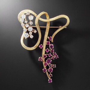 Puede incluir: Broche dorado con acabado texturizado, con un diseño estilizado. El broche está adornado con grupos de gemas redondas y transparentes y gemas redondas de color rojo intenso. El diseño tiene una forma orgánica y fluida.