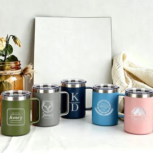 May include: A collection of five insulated travel mugs in various colors, each with a silver lid and handle. The mugs are olive green, gray, navy blue, light blue, and pink. Each mug has a unique engraved design or text.
