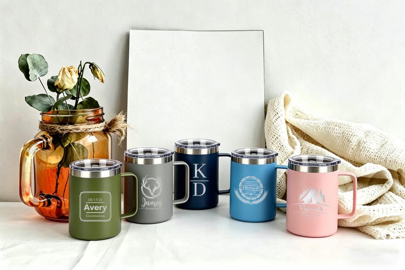 May include: A collection of five insulated travel mugs in various colors, each with a silver lid and handle. The mugs are olive green, gray, navy blue, light blue, and pink. Each mug has a unique engraved design or text.
