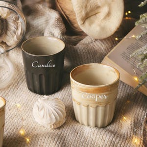 Puede incluir: Dos tazas de cerámica, una gris oscuro con "Candice" y otra crema con "Cooper" inscritos. Las tazas tienen un diseño acanalado y una apariencia rústica y artesanal. También se ven un malvavisco decorativo y luces de hadas.
