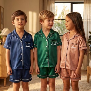 Può includere: Tre bambini con pigiami in raso coordinati. I completi includono una camicia con bottoni e pantaloncini, ciascuno con finiture bianche. I pigiami sono blu navy, verde smeraldo e rosa cipria. Ogni camicia ha un nome ricamato sulla tasca.