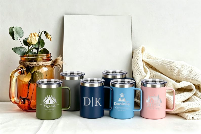 May include: A collection of insulated travel mugs in various colors, including green, navy, blue, and pink. Each mug features a silver lid and handle, with personalized text and graphics. A decorative jar with flowers is in the background.