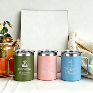 May include: Three insulated travel mugs in olive green, pink, and light blue. Each mug has a silver lid and handle. The olive green mug has a graphic of a person in a hat and the name "Danielle". The pink mug has a whale graphic. The blue mug has a golf graphic.