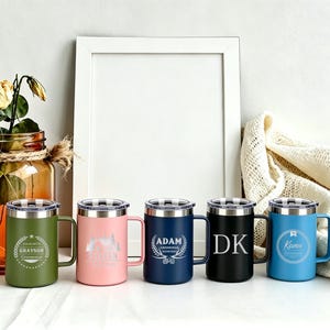 May include: Five stainless steel travel mugs in olive green, pink, navy blue, black, and light blue, each with a silver lid and handle. The mugs have personalized engravings, including names and designs. A decorative jar with flowers is in the background.