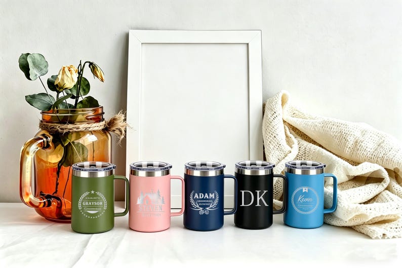 May include: Five stainless steel travel mugs in olive green, pink, navy blue, black, and light blue, each with a silver lid and handle. The mugs have personalized engravings, including names and designs. A decorative jar with flowers is in the background.