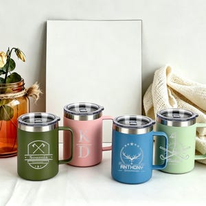 May include: Four insulated travel mugs in green, pink, blue, and light green, each with a silver lid and handle. The green mug has an axe design and the name "Soumia". The pink mug has the initials "KD". The blue mug has a deer design and the name "Anthony". The light green mug has a golf club design.