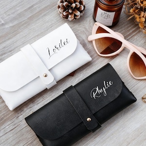 May include: Two leatherette glasses cases, one white with "Lorilei" and one black with "Rhylie" in script. A pair of pink sunglasses and gold rings are also visible. The cases are on a wooden surface.
