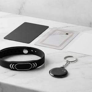 May include: A black wristband, a black key fob, and three rectangular cards are displayed on a white marble surface. The wristband has a silver accent and a black oval. The key fob is round with a silver keyring.