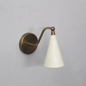 Op de afbeelding: Een wandlamp met een messing afwerking en een conische, crèmekleurige kap. De armatuur heeft een gebogen arm en een ronde basis, wat een vintage esthetiek creëert. De kap is ongeveer 15 cm hoog.