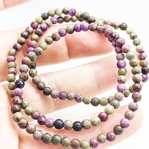 May include: Three strands of beaded bracelets. The beads are small and round, in shades of purple, green, and brown. The bracelets are held in a hand against a white background. The beads have a natural, organic appearance.
