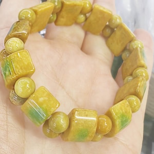 Op de afbeelding: Een armband met een mix van geelgroene, rechthoekige en bolvormige kralen. De armband heeft een mix van kleuren, waaronder geel, groen en bruin. De kralen zijn aan elkaar geregen, waardoor een uniek sieraad ontstaat.