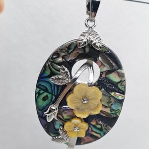 Può includere: Ciondolo ovale con baccello argentato e decorazioni floreali. Il ciondolo presenta una base di conchiglia di abalone iridescente e scintillante con un design floreale argentato e due fiori gialli. Il ciondolo è sospeso a un sottile cordino nero.