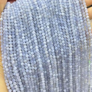 May include: Close-up of strands of light blue faceted gemstone beads. The beads are small and round, with a sparkling, polished finish. The beads are strung together, creating a flowing, textured appearance. The overall color is a soft, pale blue.
