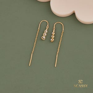 以下が含まれることがあります： 小さな輝く宝石が付いた、ゴールドカラーのスレッドピアス。ピアスは繊細なチェーンと、端にまっすぐなバーが付いています。緑色の表面に展示されています。