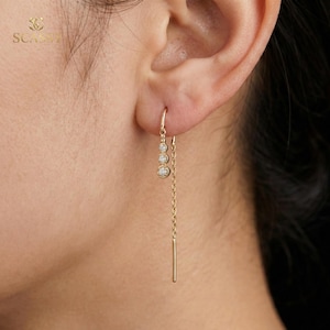 Puede incluir: Un pendiente dorado con tres pequeñas piedras preciosas redondas y transparentes y una cadena colgante con una barra rectangular en el extremo. El pendiente se lleva en la oreja de una persona.