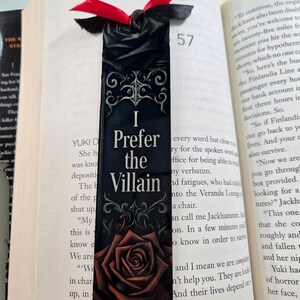 Może przedstawiać: Czarna zakładka do książki z napisem "I Prefer the Villain" i ilustracją czerwonej r&oacute;ży. Zakładka ma przymocowaną czarną i czerwoną wstążkę i jest umieszczona w otwartej książce.