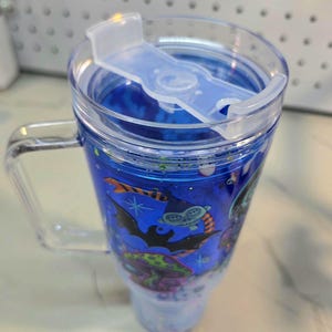 May include: A clear plastic tumbler with a handle and a lid featuring a sliding closure. The tumbler contains a blue liquid with floating glitter and a graphic print of a green creature, a black bat, and a colorful mushroom on the exterior.