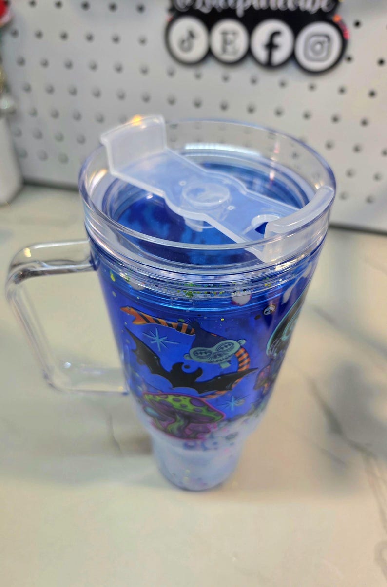 May include: A clear plastic tumbler with a handle and a lid featuring a sliding closure. The tumbler contains a blue liquid with floating glitter and a graphic print of a green creature, a black bat, and a colorful mushroom on the exterior.